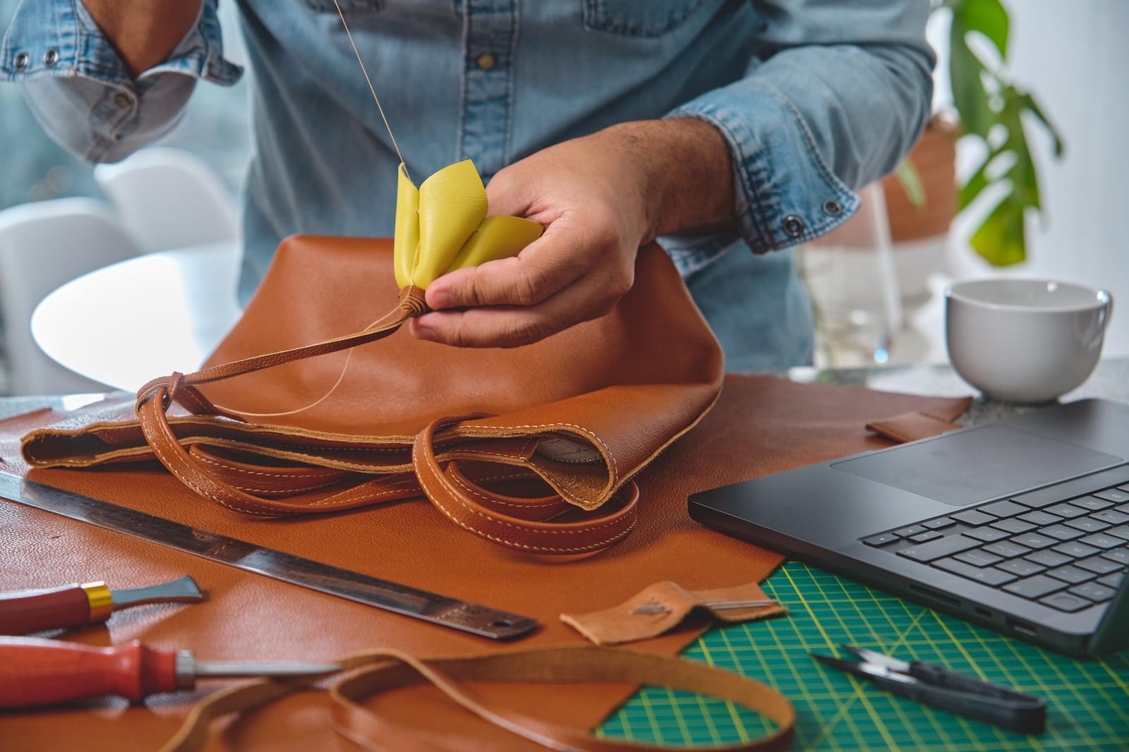 Artesão costurando à mão a alça de uma bolsa de couro caramelo na bancada de trabalho